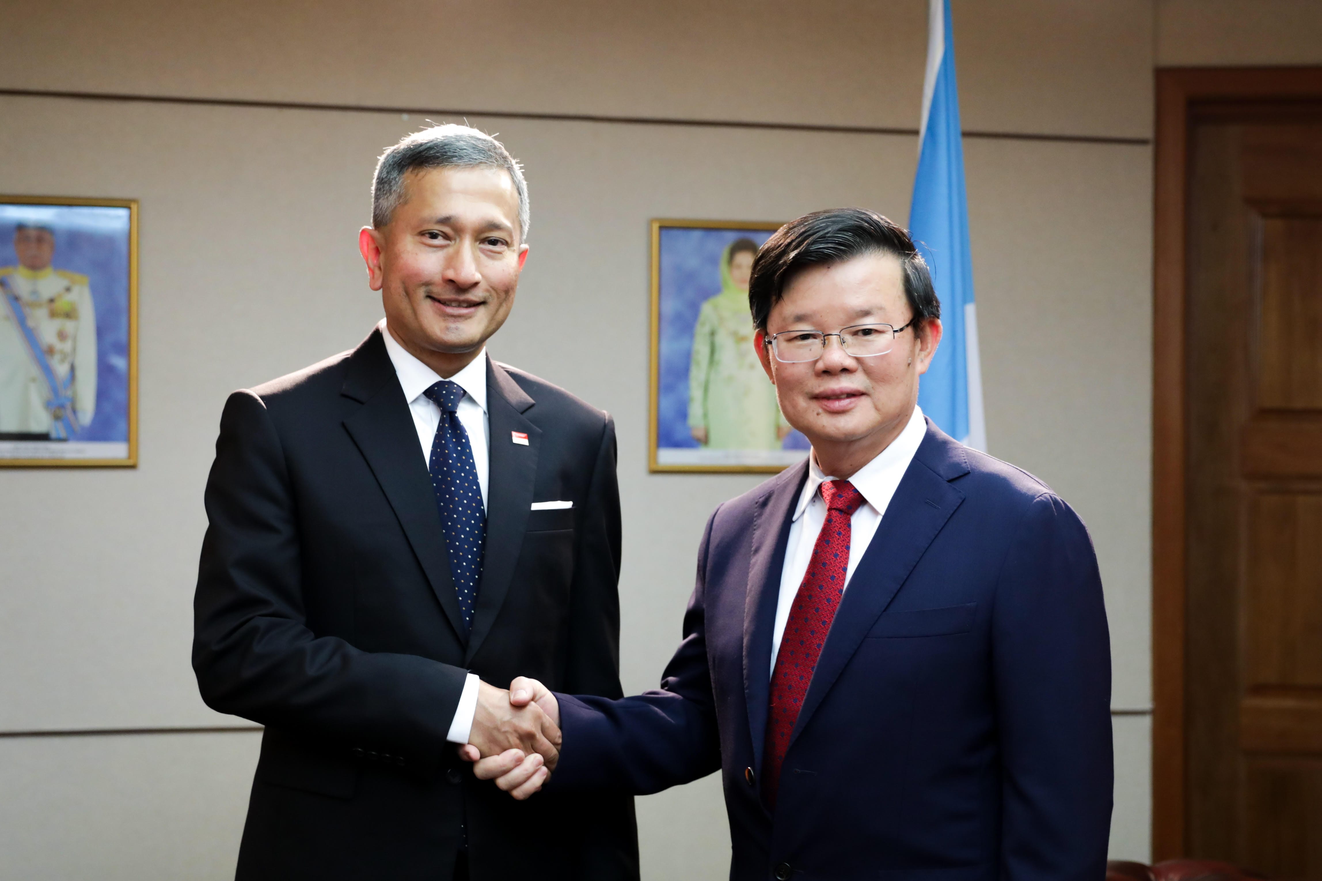 Two men in suits shake hands; Singaporean flag pin on one, flags and portraits behind.